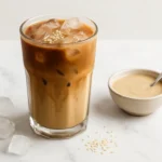 Glass of tahini iced latte on marble surface with ice cubes, sesame seeds, and a bowl of tahini beside it