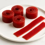 A close-up of vibrant red zero-calorie fruit roll-ups, both spiraled and flat, neatly arranged on a white round plate over a marble background.